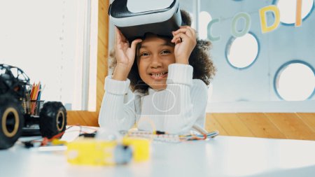 Photo for African girl taking off VR glass or head set while looking at camera. Student smiling at camera with colored pencil and laptop placed on table in STEM technology class. Online education. Erudition. - Royalty Free Image