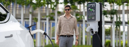 Téléchargez les photos : Jeune homme recharger la batterie des voitures électriques de la station de charge dans le parc urbain vert extérieur. Voiture rechargeable pour des déplacements urbains respectueux de l'environnement. Panorama Expedient - en image libre de droit