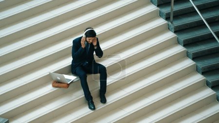 Téléchargez les photos : Détente homme d'affaires intelligent écoutant de la musique du casque tout en étant assis à l'escalier. Happy manager souriant en dansant ou en passant à la musique. Investisseur recevoir de bonnes nouvelles, obtenir une promotion. Exultant. - en image libre de droit