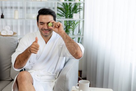 Photo for Man holding slices of fresh cucumber and wearing bathrobe enjoying luxurious facial skincare spa in resort or hotel. Skin treatment for face and beauty care. Quiescent - Royalty Free Image