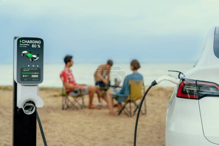 Photo for Alternative family vacation trip traveling by the beach with electric car recharging battery from EV charging station with blurred family enjoying the seascape background. Perpetual - Royalty Free Image