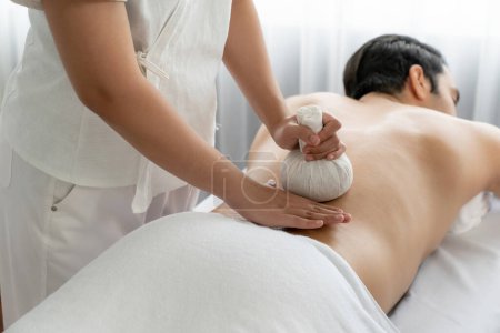 Photo for Hot herbal ball spa massage body treatment, masseur gently compresses herb bag on man body. Tranquil and serenity of aromatherapy recreation in day lighting ambient at spa salon. Quiescent - Royalty Free Image