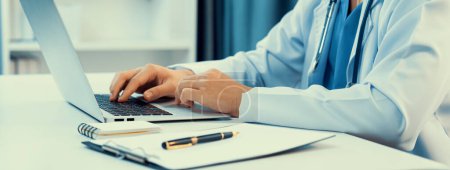 Photo for Doctor at hospital sit at his desk working on laptop diagnosing patient test results, developing treatment plan for illnesses and sicknesses. Medical staff and healthcare service. Panorama Rigid - Royalty Free Image