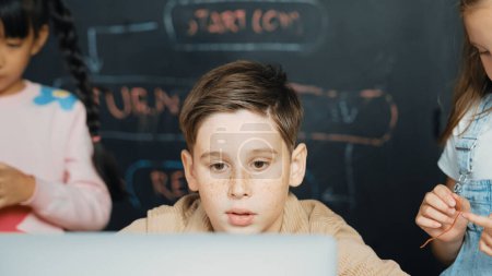 Photo for Closeup of boy using laptop programing engineering code and writing program while group of diverse kid holding controller in STEM technology classroom at blackboard written with prompt. Erudition. - Royalty Free Image
