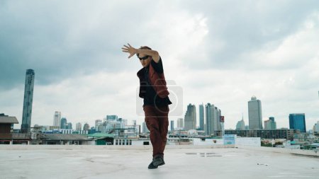 Téléchargez les photos : Tournage en mouvement de la performance de danse B-boy par un danseur de rue professionnel sur le toit avec gratte-ciel, vue sur la ville. Attrayant hipster asiatique montrer pas énergique. Sport de plein air 2024. Endeavor. - en image libre de droit
