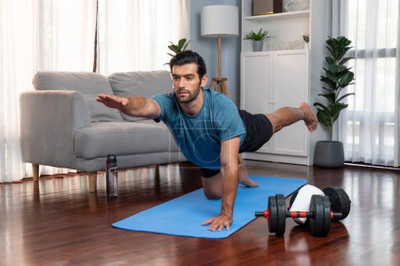 Téléchargez les photos : Homme flexible et dextérité en tenue de sport faisant position de yoga en posture de méditation sur tapis d'exercice à la maison. Mode de vie sain de yoga de gaieté à domicile avec l'esprit paisible et la sérénité. - en image libre de droit