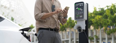 Photo for Young man use smartphone to pay for electricity at public EV car charging station green city park. Modern environmental and sustainable urban lifestyle with EV vehicle. Panorama Expedient - Royalty Free Image
