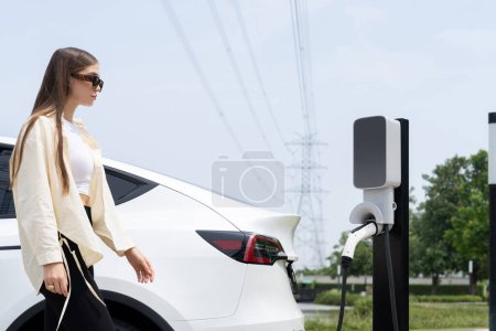 Téléchargez les photos : Jeune femme recharger la batterie de voiture EV à la station de charge connectée à l'installation industrielle électrique de tour de réseau électrique comme industrie électrique pour l'utilisation de véhicule respectueuse de l'environnement. Expedient - en image libre de droit