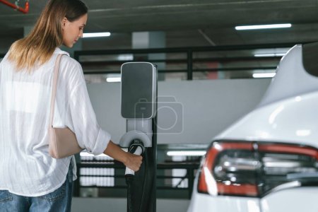Téléchargez les photos : Jeune femme Voyage avec EV voiture électrique au centre commercial parking charge dans le centre-ville montrant mode de vie durable urbain par vert propre énergie rechargeable de véhicules électriques à l'intérieur - en image libre de droit