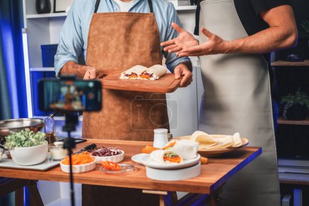 Photo for Cropped view of chef influencers presenting fresh salad roll on cooking step by step, streaming via smartphone on social media live channel, showing ingredients vegetable soft tortilla wrap. Sellable. - Royalty Free Image
