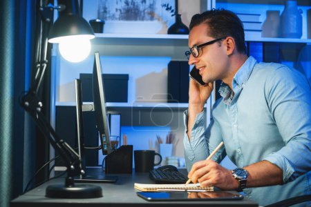 Photo for Working businessman calling phone with customer, typing brief message at neon workplace, planning online business using information project at night time surrounded by stationary on desk. Sellable. - Royalty Free Image