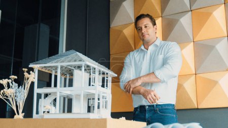 Photo for Portrait of smiling confident architect engineer looking at camera while standing with arm folded with house model placed on table. Professional businessman crossing arm. Design concept. Manipulator. - Royalty Free Image