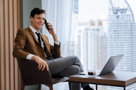 Photo for Closeup of handsome businessman making phone call with manager while sitting near window with skyscraper view. Executive manager talking working by using phone and laptop. Look aside. Ornamented. - Royalty Free Image
