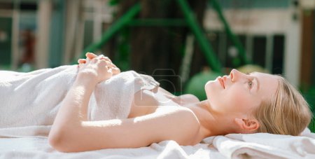Téléchargez les photos : Portrait de belle fille caucasienne en serviette blanche se trouve sur le lit de spa paisiblement à l'extérieur entouré d'un environnement relaxant et apaisant en attendant le massage du corps. Fond flou. Tranquillité - en image libre de droit