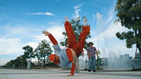 Téléchargez les photos : Asiatique hipster habile faisant pose de gel à la caméra entourée par l'équipe de danseur de rupture près de la fontaine avec fond de ciel. Groupe de belles personnes pratiquent la danse b-boy. Sport de plein air 2024. Brillamment. - en image libre de droit