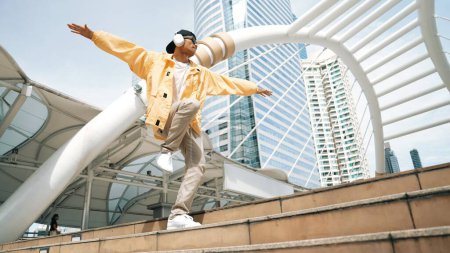 Photo for Stylish asian man perform hiphop dancing with low angle camera. Break dancer walking up the stair while listening music from headphone and dancing together at city. Outdoor sport 2024. Sprightly. - Royalty Free Image