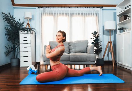 Téléchargez les photos : Femme flexible et dextérité en tenue de sport faisant position de yoga en posture de méditation sur tapis d'exercice à la maison. Mode de vie sain de yoga de gaieté à domicile avec l'esprit paisible et la sérénité. - en image libre de droit