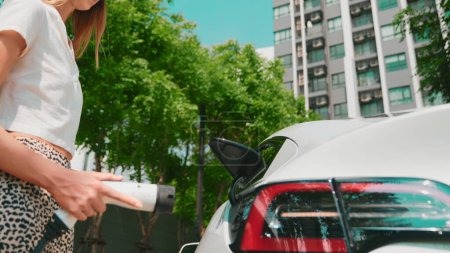 Téléchargez les photos : Jeune femme recharger la batterie de voiture électrique EV dans la zone résidentielle borne de recharge EV dans la ville urbaine condo avec un mode de vie durable vert et renouvelable d'énergie propre pour les entrailles de véhicules électriques - en image libre de droit