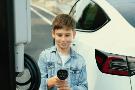 Photo for Road trip vacation with eco-friendly EV car and little boy pointing EV charger at camera. Electric vehicle and charging station using clean and sustainable energy for environment protection. Perpetual - Royalty Free Image