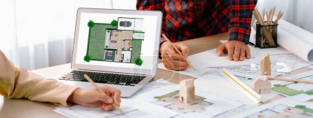 Photo for Professional architect represented ECO house displayed on laptop to project manager on messy table with wooden block, blueprint, architectural document placed at modern office. Close up. Delineation. - Royalty Free Image