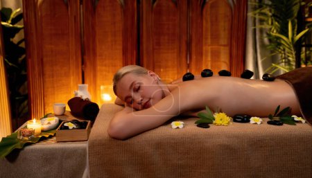 Téléchargez les photos : Massage aux pierres chaudes au salon de spa dans un complexe de luxe avec lumière chaude à la bougie, une cliente heureuse profitant d'un massage aux pierres de basalte spa glisse sur le corps avec une chaleur apaisante. Tranquillité - en image libre de droit