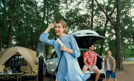 Photo for Outdoor adventure and family vacation camping at sea travel by eco friendly car. Cheerful woman or mother holding, pointing EV charger point with playful and happiness posture in campsite. Perpetual - Royalty Free Image