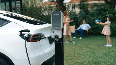Photo for Electric vehicle recharge from home charging station on background of happy and playful family playing together. EV car using alternative and sustainable energy for better future Synchronos - Royalty Free Image