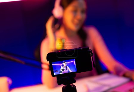 Téléchargez les photos : Écran de caméra de mise au point sélective dans le canal hôte de belle asiatique écoutant la musique sur les médias sociaux avec l'auditeur, portant des écouteurs pastel sur fond flou en utilisant micro au studio néon. Stratagème. - en image libre de droit