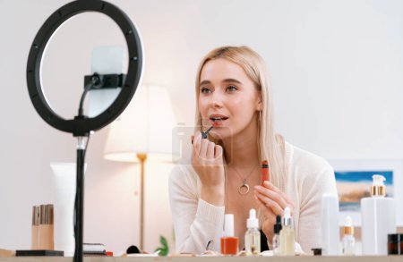 Photo for Young woman making beauty and cosmetic tutorial video content for social media. Beauty blogger using camera and light ring while showing how to apply liquid lipstick to audience or follower. Blithe - Royalty Free Image