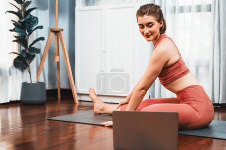 Téléchargez les photos : Femme flexible et dextérité en tenue de sport faisant position de yoga en posture de méditation sur tapis d'exercice à la maison. Gaieté saine séance de formation en ligne de yoga à domicile avec l'esprit paisible et la sérénité. - en image libre de droit