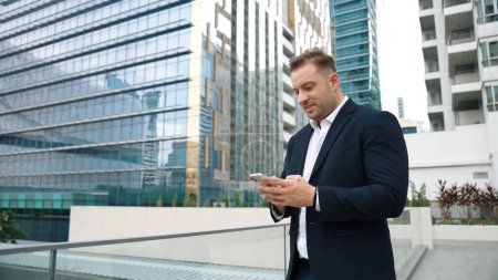 Photo for Happy businessman using phone to chatting with team while standing at urban city. Front view of project manager working on telephone with skyscraper view while communicate with marketing team. Urbane. - Royalty Free Image
