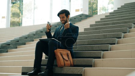 Photo for Successful business man celebrate successful project while sitting at stairs. Smart project manager getting new gob, getting promotion, increasing sales while calling friends by using phone. Exultant. - Royalty Free Image