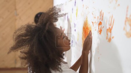 Téléchargez les photos : Africaine fille peindre le mur avec des mains colorées teintées avec un ami. Groupe d'apprenants multiculturels debout devant le mur tout en utilisant la main pour créer des œuvres d'art. Concept d'activité créative. Erudition. - en image libre de droit