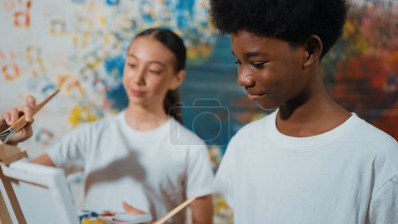 Foto de Diversos niños felices pintan lienzo junto con coloridas paredes manchadas. Estudiante multicultural inteligente dibujando juntos en la lección de arte mientras usa camisa blanca. Concepto de actividad creativa. Edificación. - Imagen libre de derechos