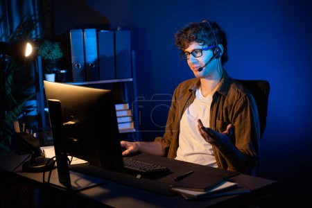 Photo for Young creative businessman communicating with listeners on laptop screen or meeting with team member, wearing headphones at neon light modern home office. Concept of working at night time. Gusher. - Royalty Free Image