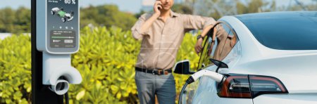Photo for Man talking on the phone while recharge EV car battery at charging station connected to power grid tower electrical as electrical industry for eco friendly car utilization. Panorama Expedient - Royalty Free Image