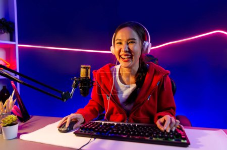 Photo for Host channel of smiling beautiful Asian girl streamer playing online game wearing headphone talking with viewers media online recording phone. Esport skilled team players in neon blue room. Stratagem. - Royalty Free Image