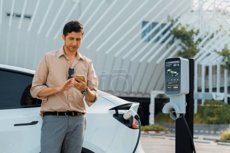 Photo for Young man use smartphone to pay for electricity at public EV car charging station at city commercial mall parking lot. Modern environmental and sustainable urban lifestyle with EV vehicle. Expedient - Royalty Free Image