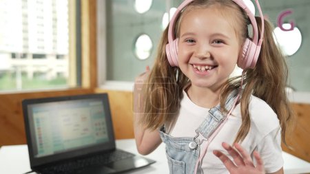 Photo for Smiling girl looking while waving hand at camera with laptop placed on table. Child wearing headphone smiling while laptop screen show system programing or coding program in STEM class. Erudition. - Royalty Free Image