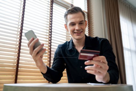 Photo for Young man sitting at table using online payment app and digital wallet on smartphone to pay with credit card. E commerce shopping and modern purchasing via mobile internet. Unveiling - Royalty Free Image