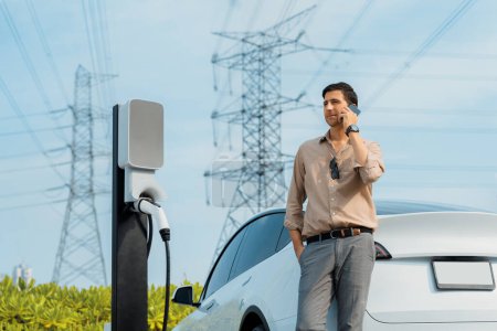 Photo for Man talking on the phone while recharge EV car battery at charging station connected to power grid tower electrical as electrical industry for eco friendly car utilization.Expedient - Royalty Free Image