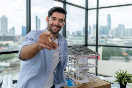 Photo for Portrait of smart engineer in casual outfit smiling at camera while holding pencil. Businessman pointing at camera and standing near house model, project plan with sky scraper behind. Tracery. - Royalty Free Image