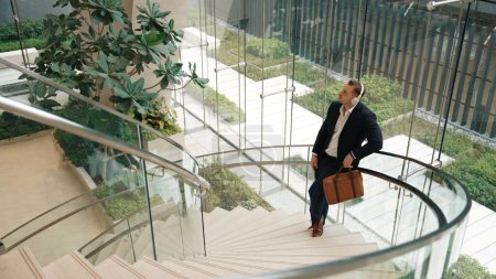 Top view of caucasian businessman listening relaxing music wearing headphone while standing at spiral stair cases at modern building. Project manager enjoy listening song while move to rhyme. Urbane.