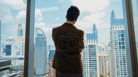 Photo for Back view ambitious businessman standing in ornamented office gazing out window to cityscape skyline. Determination and business ambition drive business career toward to bright future - Royalty Free Image