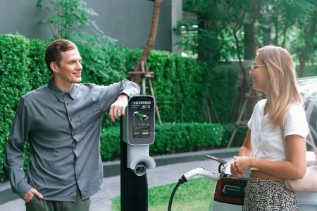 Junges Paar reist mit Elektroauto-Aufladung im grünen nachhaltigen Stadtgarten im Sommer zeigt urbanen Nachhaltigkeitslebensstil durch grüne saubere wiederaufladbare Energie von Elektrofahrzeuginnern