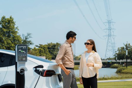 Photo for Young couple recharge EV car battery at charging station connected to power grid tower electrical industrial facility as electrical industry for eco friendly vehicle utilization. Expedient - Royalty Free Image