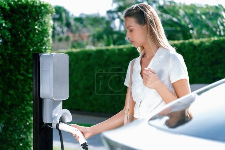 Téléchargez les photos : Les jeunes femmes voyagent avec la voiture électrique EV charge dans le jardin vert durable de la ville en plein air en été. Style de vie urbain durable par vert propre énergie rechargeable des véhicules électriques BEV à l'intérieur - en image libre de droit