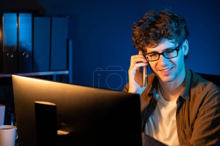 Photo for Young businessman calling on smartphone to customer or coworker, looking at computer screen of creative presentation surround work desk at neon modern office. Concept of new next project. Gusher. - Royalty Free Image