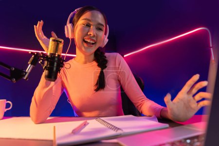 Photo for Smiling creative young beautiful Asian influencer talking in channel with positive podcast speech at night time, using laptop for presentation online at pink neon light modern studio. Stratagem. - Royalty Free Image