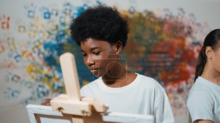 Photo for African boy painting canvas with watercolor while happy caucasian girl draw cool tone picture at colorful stained wall. Multicultural highschool student attend creative activity together. Edification. - Royalty Free Image